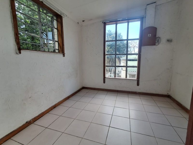 An empty bedroom featuring tiled flooring, timber-framed windows with security bars, and an exposed electrical distribution board, requiring cosmetic renovation.