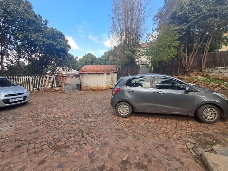 A paved driveway area featuring a brick boundary wall, metal security gate, and mature trees, providing access to the property entrance.