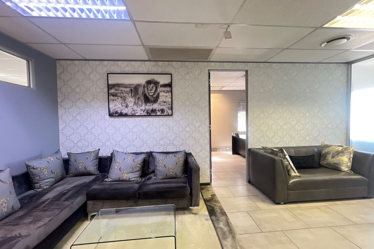 Commercial office lounge area featuring tiled flooring, suspended ceiling with integrated lighting, and a glass partition leading to an adjacent workspace.