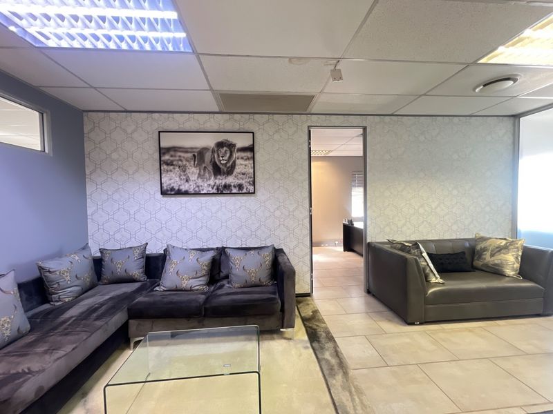 Commercial office lounge area featuring tiled flooring, suspended ceiling with integrated lighting, and a glass partition leading to an adjacent workspace.