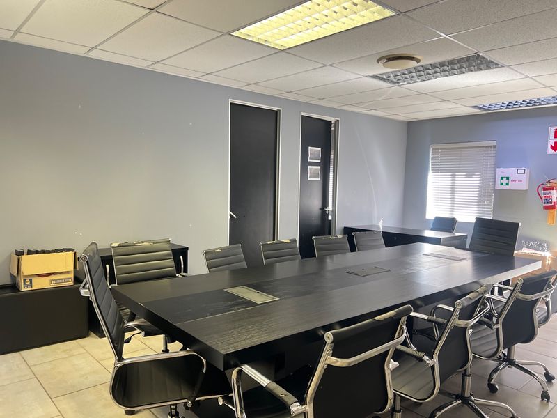 A functional commercial boardroom featuring a large central table, suspended ceiling with recessed lighting, tiled flooring, and window blinds.