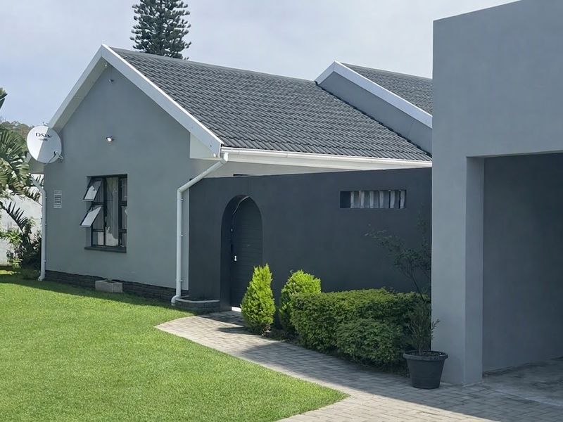 A well-maintained modern house exterior featuring a grey facade, tiled roof, paved walkway, and a private arched entrance gate set within a landscaped garden.