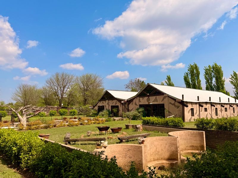 A view of the rear garden featuring a large barn-style building with a metal roof, landscaped grounds, and built-in masonry seating areas.