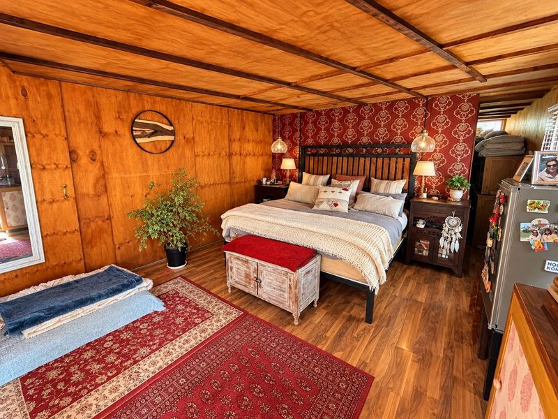 A rustic bedroom featuring warm wood-paneled walls, exposed ceiling beams, and a combination of patterned wallpaper and laminate flooring.