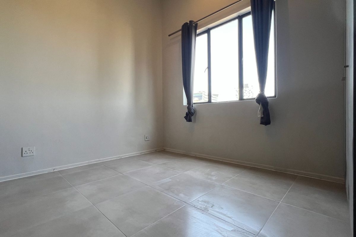 A bright, empty bedroom featuring neutral tiled flooring and a large window, providing a clean and versatile space ready for furnishing.