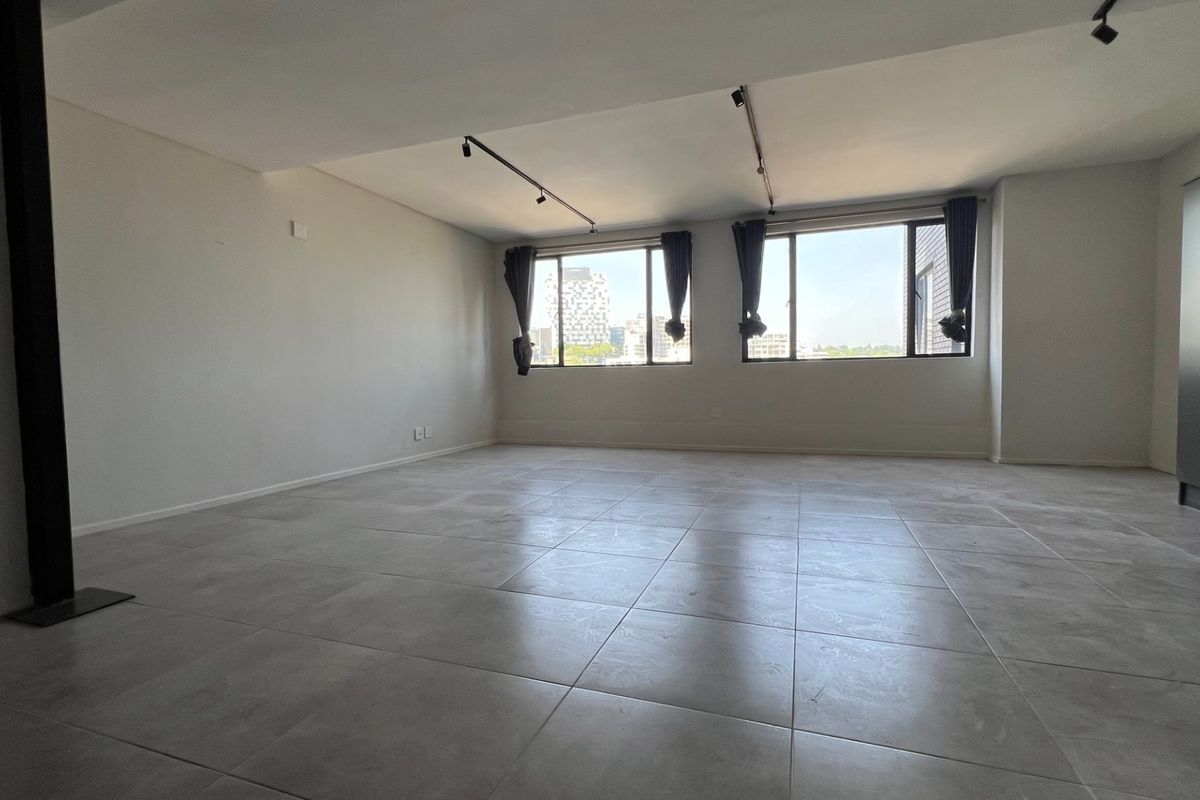 A spacious, empty living area featuring neutral tiled flooring, modern track lighting, and large windows providing natural light and urban views.