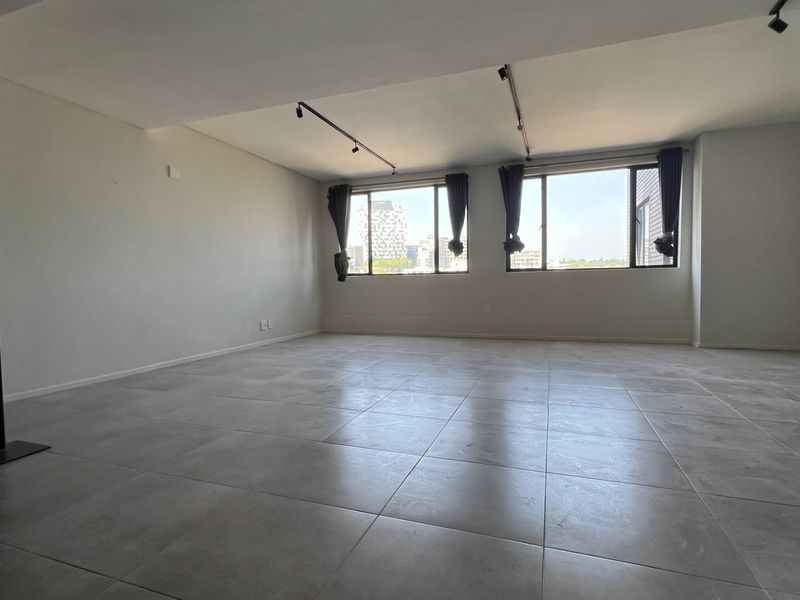 A spacious, empty living area featuring neutral tiled flooring, modern track lighting, and large windows providing natural light and urban views.