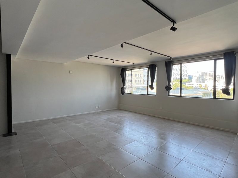 Spacious, light-filled living area featuring neutral tiled flooring, modern track lighting, and large windows offering an urban outlook.