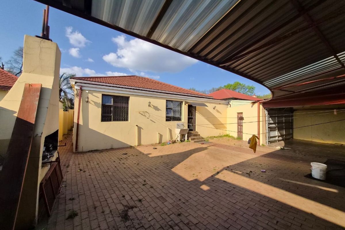 Rear exterior view featuring a paved courtyard, built-in braai, covered parking area, and utility sink against the main house structure.