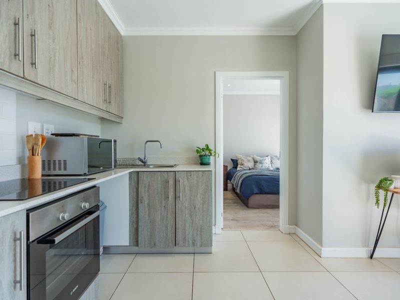 Modern kitchen featuring light wood-grain cabinetry, stone countertops, integrated oven and hob, and neutral tiled flooring, with a view into the adjacent bedroom.