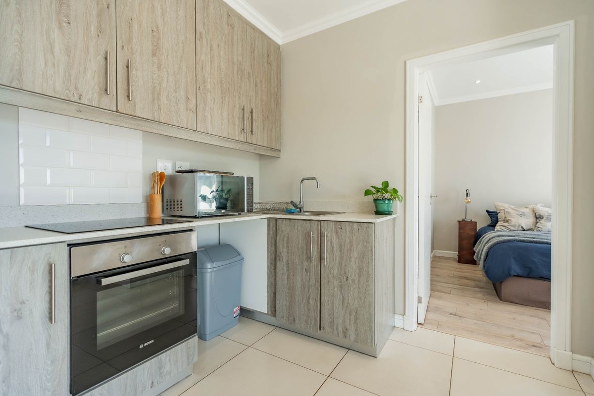 A compact, modern kitchen featuring wood-look cabinetry, built-in oven and hob, subway tile backsplash, and tiled flooring, with an adjacent bedroom view.
