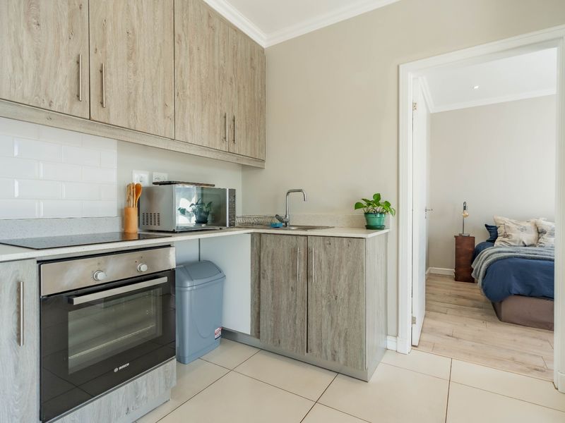 A compact, modern kitchen featuring wood-look cabinetry, built-in oven and hob, subway tile backsplash, and tiled flooring, with an adjacent bedroom view.