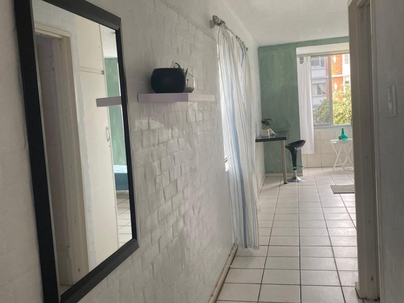 A narrow hallway featuring white painted brick walls and tiled flooring, leading towards a sunlit area with a window.