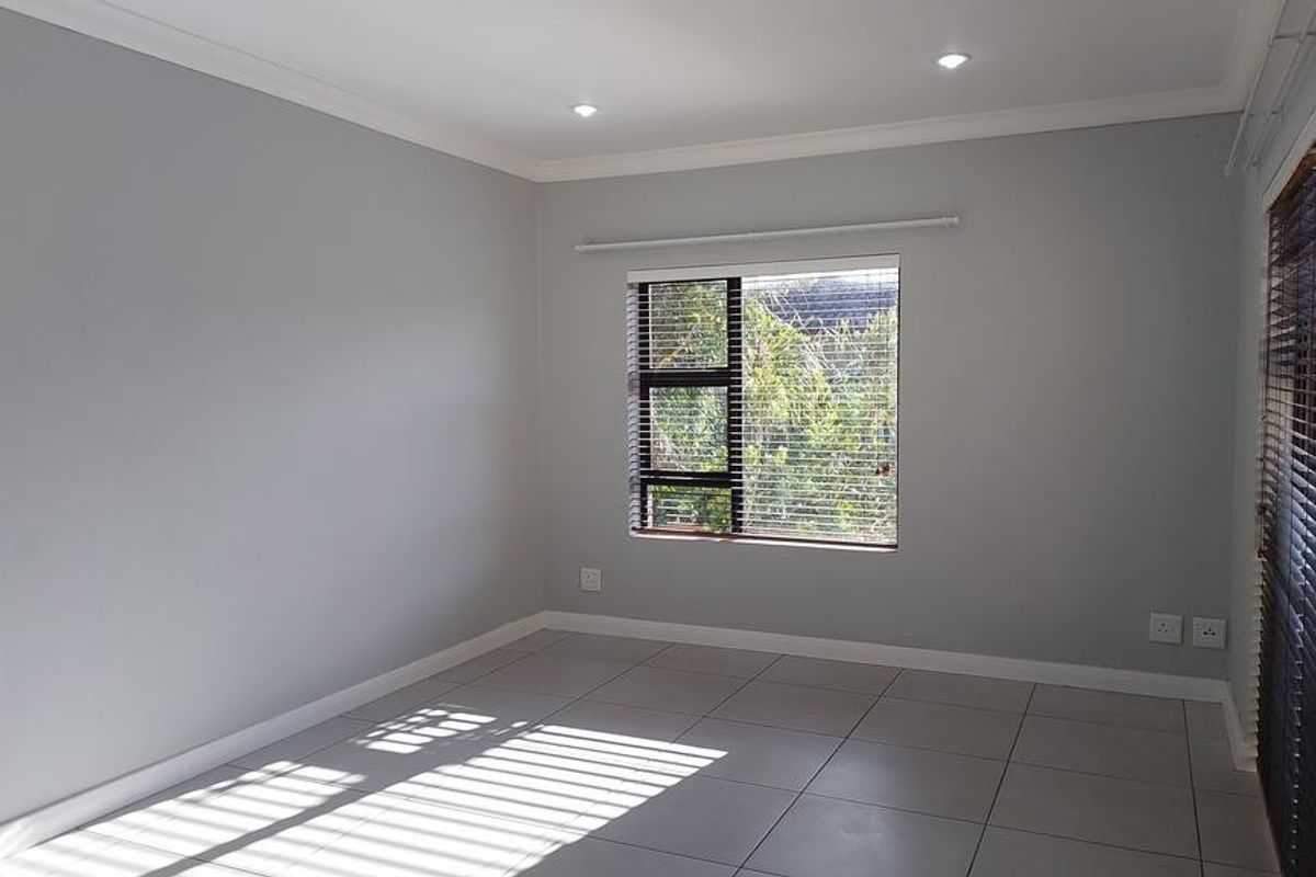 A bright, empty living area featuring neutral grey walls, large tiled flooring, recessed downlights, and access to an outdoor space through a sliding door.