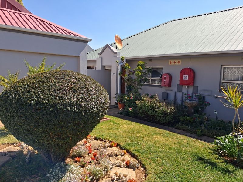 A well-maintained exterior rear view of a retirement village unit featuring a landscaped garden, corrugated metal roofing, and installed fire safety equipment.