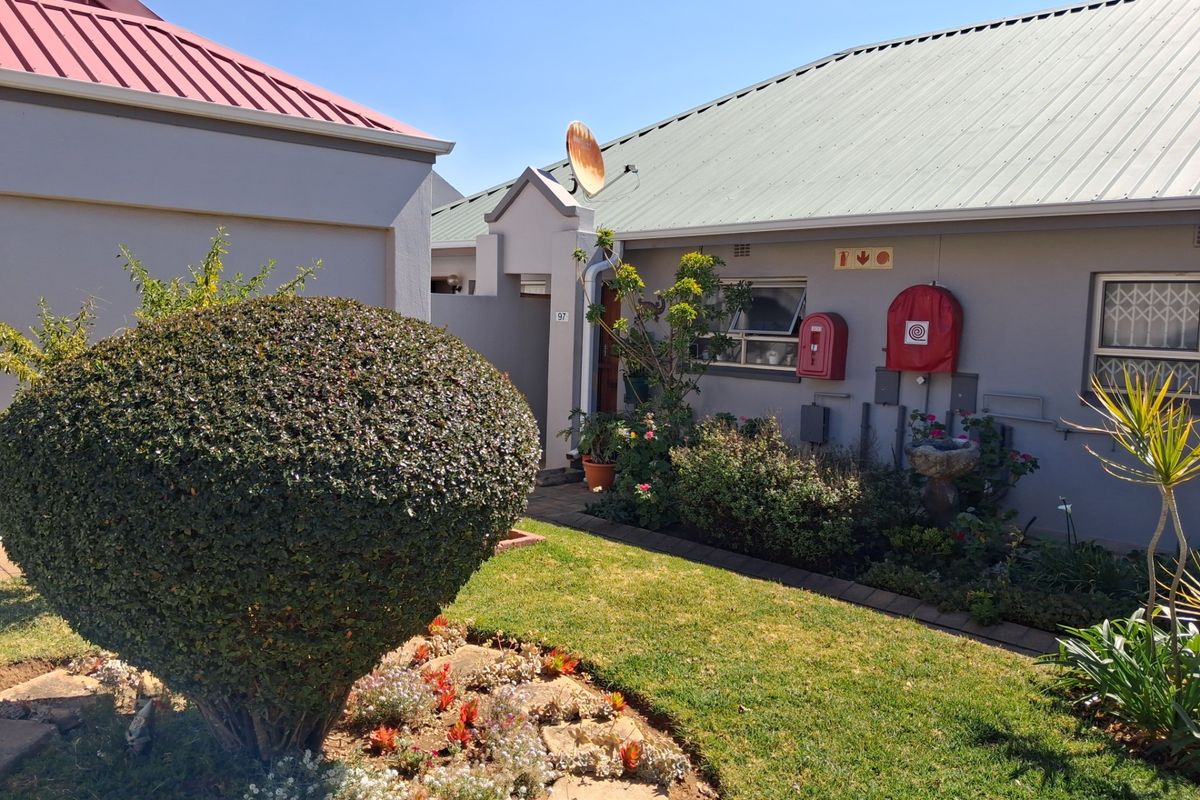 A well-maintained exterior rear view of a retirement village unit featuring a landscaped garden, corrugated metal roofing, and installed fire safety equipment.