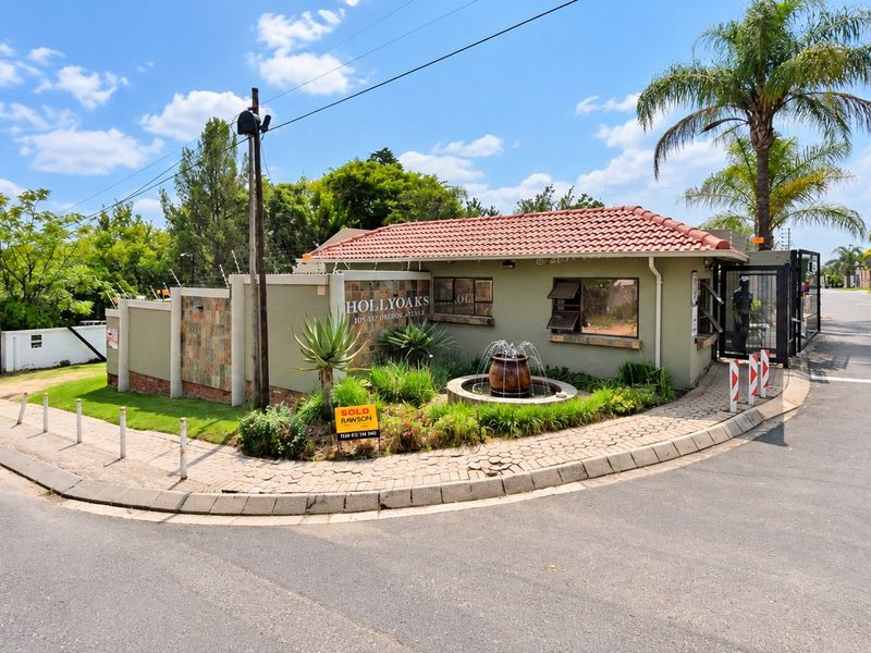 Front entrance and security gatehouse of the Hollyoaks residential complex, featuring a landscaped garden, water feature, and perimeter security fencing.