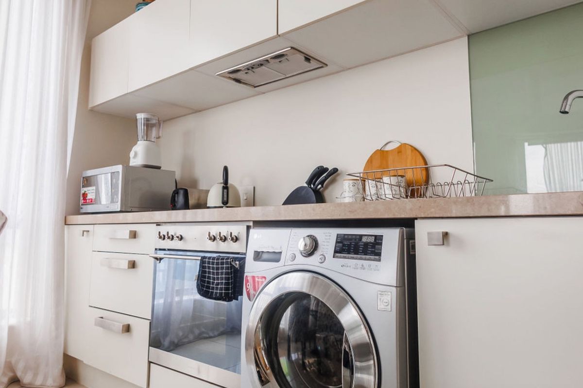 A modern, compact kitchen featuring white cabinetry, stone countertops, a built-in oven, and an integrated washing machine.