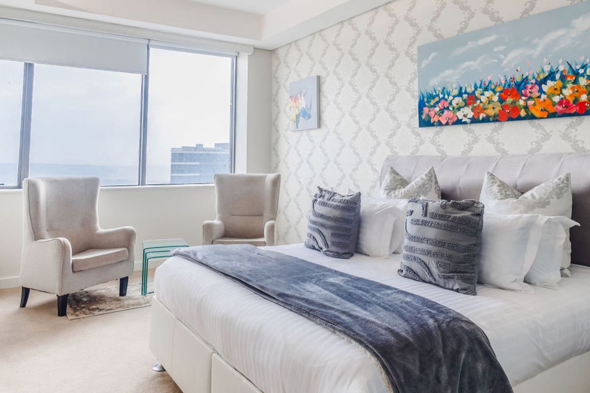 A well-lit bedroom featuring patterned wallpaper, a large window with roller blinds, and a recessed ceiling design, offering a comfortable and bright living space.