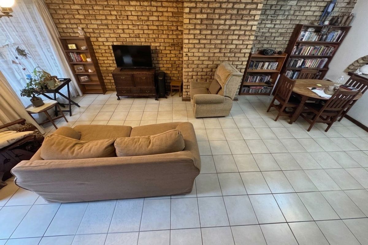 A living room featuring an exposed brick feature wall and neutral ceramic floor tiles, offering a functional and dated living space.