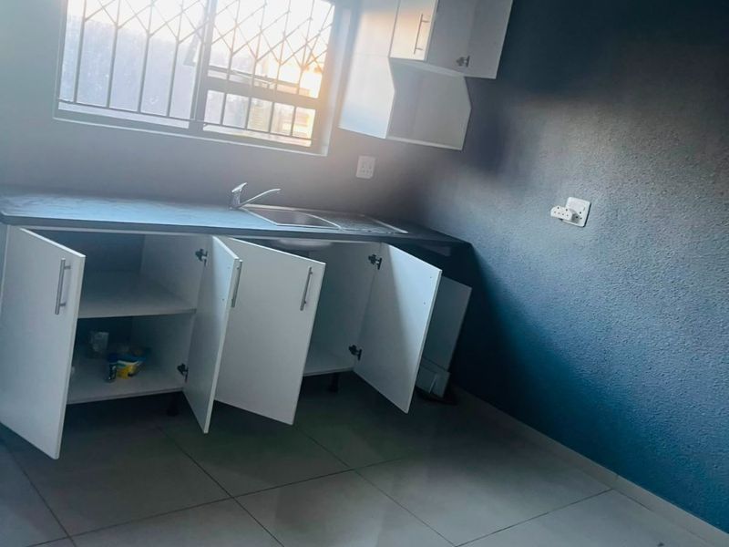 A compact kitchen area featuring white built-in cabinetry, a stainless steel sink, and tiled flooring, with security bars installed on the window.