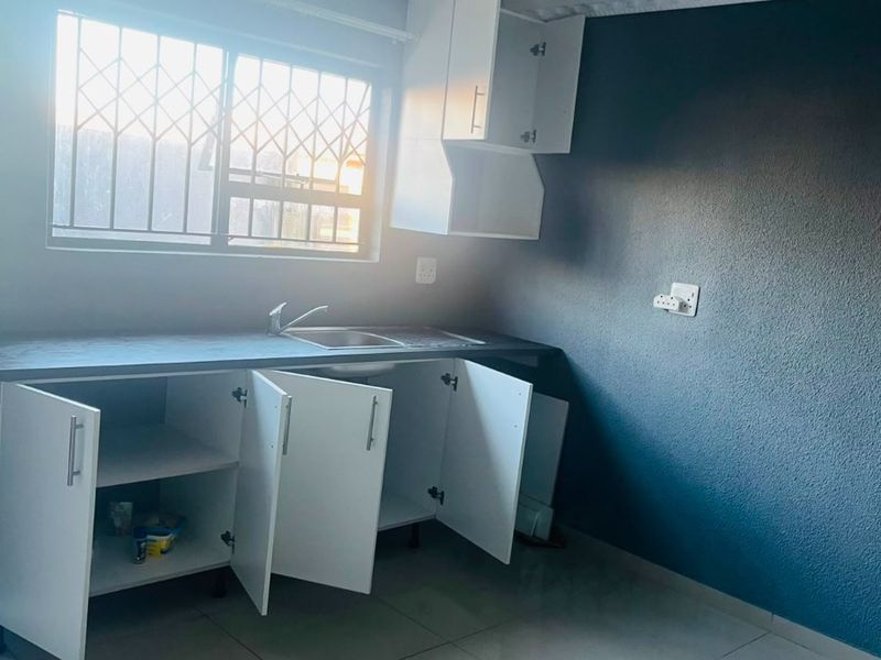 A compact kitchen area featuring white built-in cabinetry, a stainless steel sink, tiled flooring, and security bars on the window.