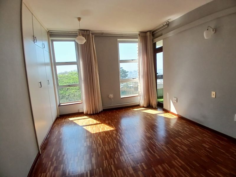 A bright, empty bedroom featuring polished parquet flooring, ample built-in cupboard space, and direct access to a balcony through a glass door.