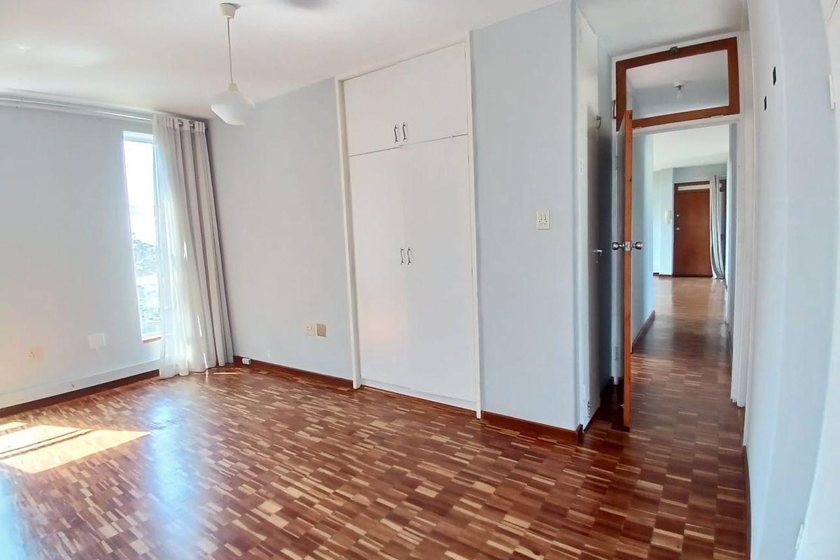 A bright, empty bedroom featuring classic parquet flooring, built-in cupboards, and a wooden door frame leading to the hallway.