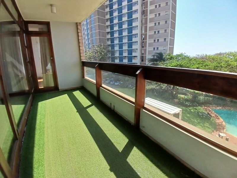 A covered balcony featuring a timber-framed glass balustrade and artificial grass flooring, offering views over the complex garden and pool area.