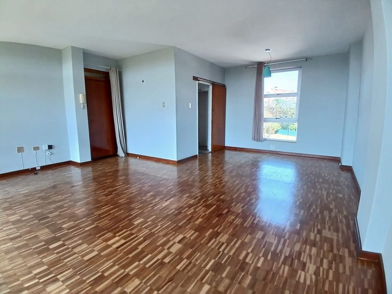A spacious, empty living room featuring classic parquet flooring, neutral walls, and natural light from a large window.
