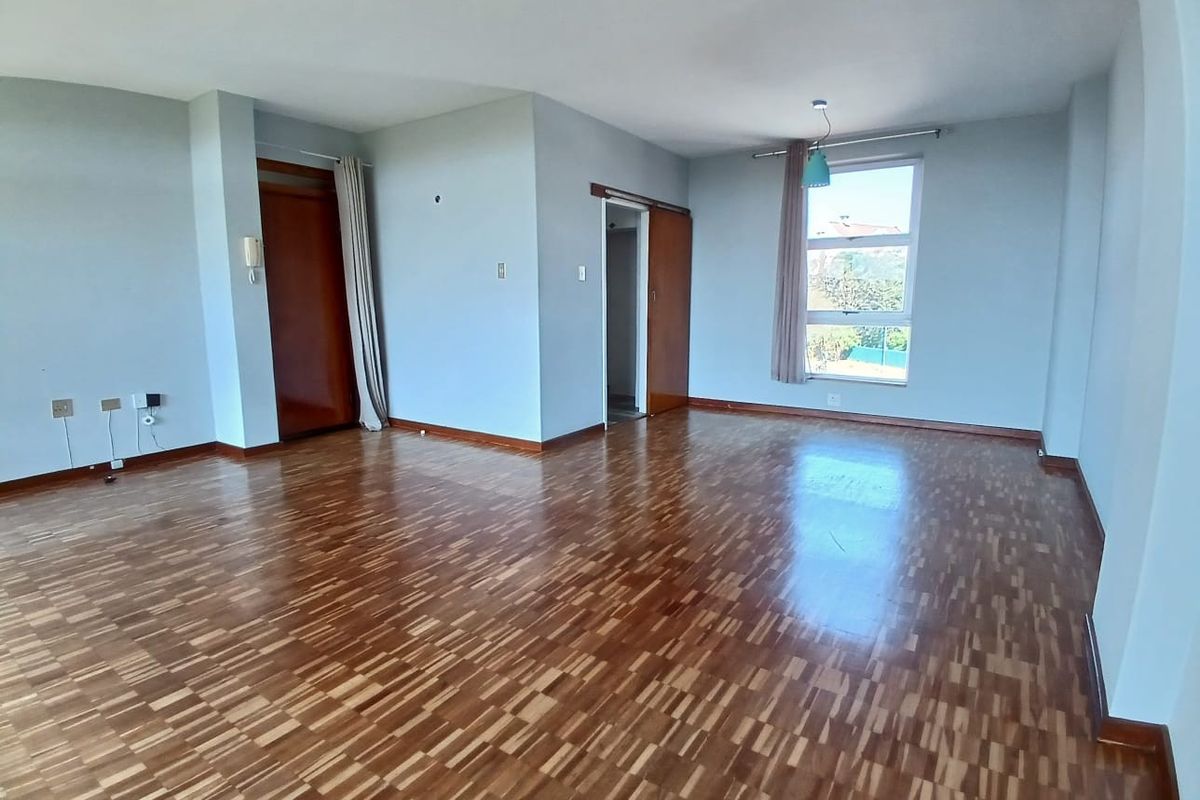 A spacious, empty living room featuring classic parquet flooring, neutral walls, and natural light from a large window.