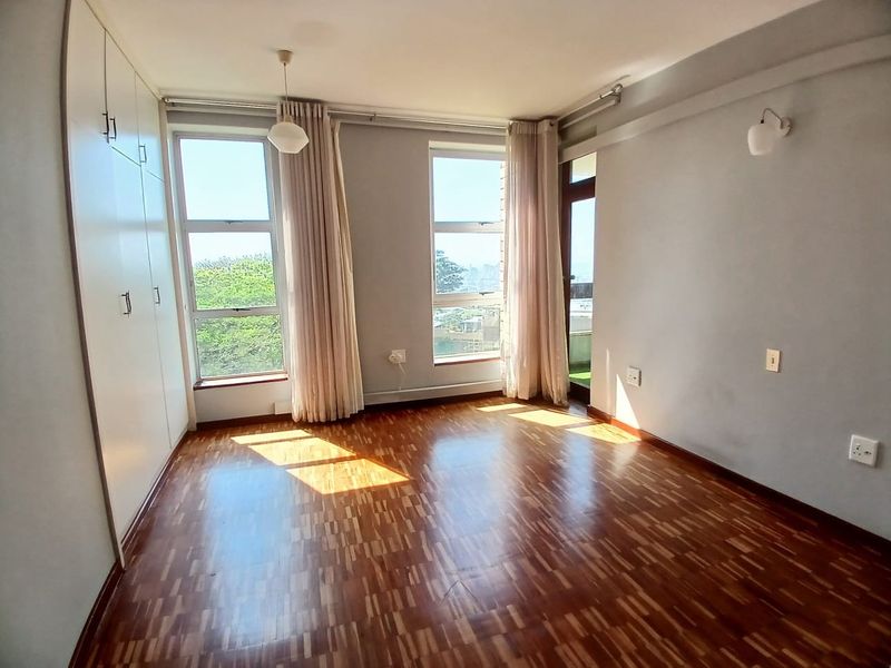 A bright, empty bedroom featuring warm parquet flooring, ample built-in cupboard space, and large windows that provide natural light and access to a balcony.