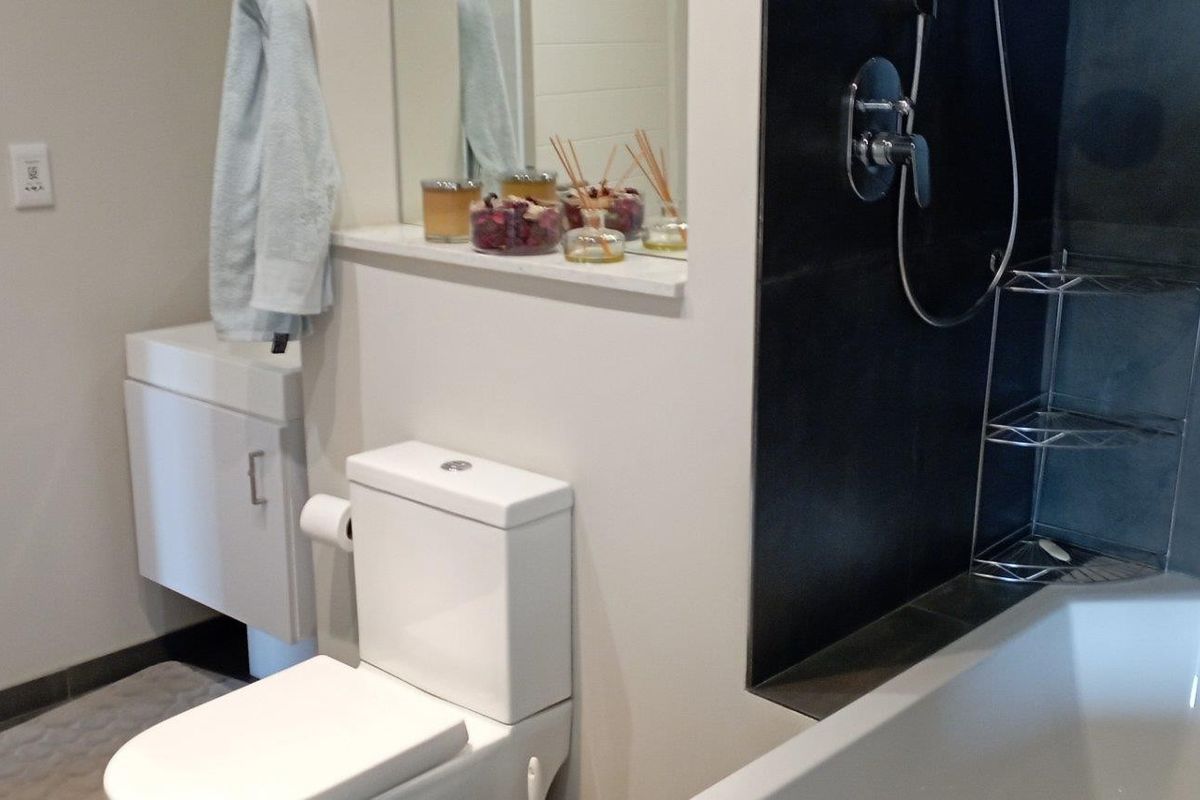 A modern bathroom featuring a white toilet, vanity unit, and a bathtub with dark tiled surround and integrated shower fixtures.
