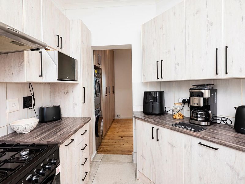 A functional scullery featuring light wood-grain cabinetry, dark laminate countertops, a gas hob with extractor, and a dedicated laundry area with stacked appliances.