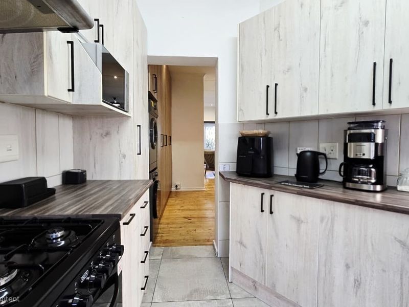 Modern galley-style kitchen featuring light wood-grain cabinetry, dark laminate countertops, a gas hob with extractor fan, and neutral tiled flooring.