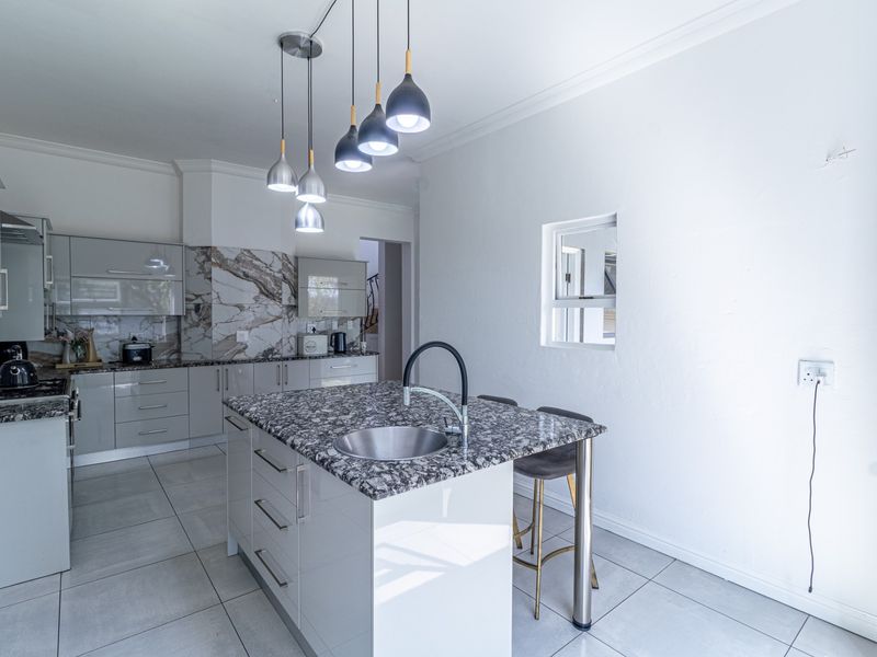Modern kitchen featuring a central island with granite countertops, undermount sink, sleek cabinetry, marble-look splashback, and contemporary pendant lighting.