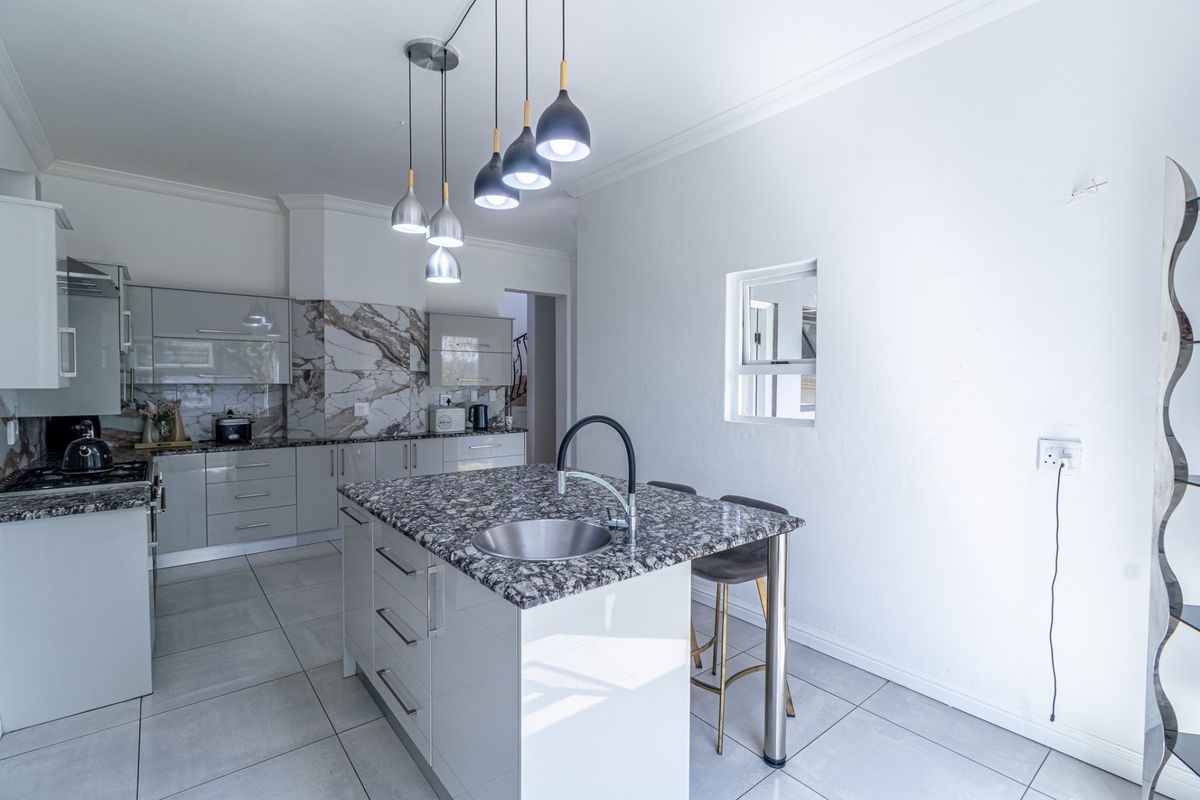 Modern kitchen featuring a central island with granite countertops, undermount sink, sleek cabinetry, marble-look splashback, and contemporary pendant lighting.