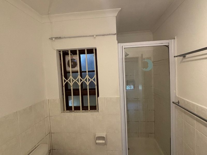 A functional bathroom featuring a shower enclosure, toilet, and vanity basin, with tiled finishes and security bars on the window.