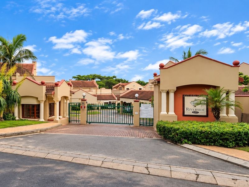 The secure gated entrance to Riverside complex, featuring a paved driveway, perimeter walls, and well-maintained landscaped gardens.