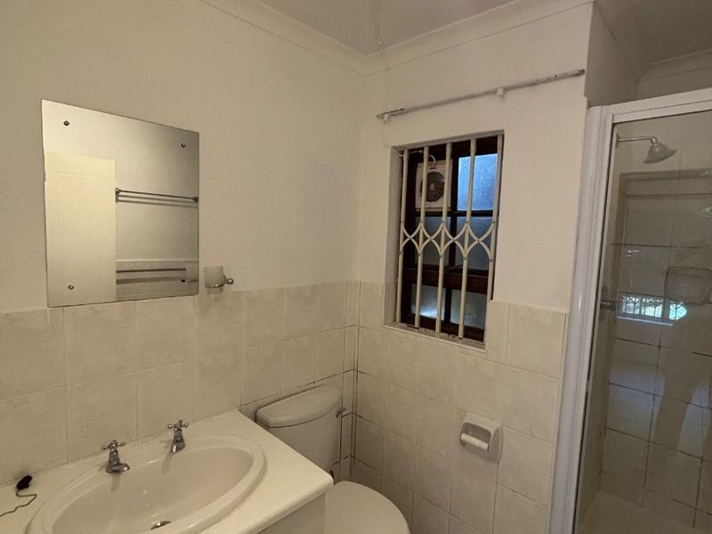 A functional bathroom featuring a vanity cabinet, toilet, and a separate shower enclosure with tiled finishes and a security-gated window.