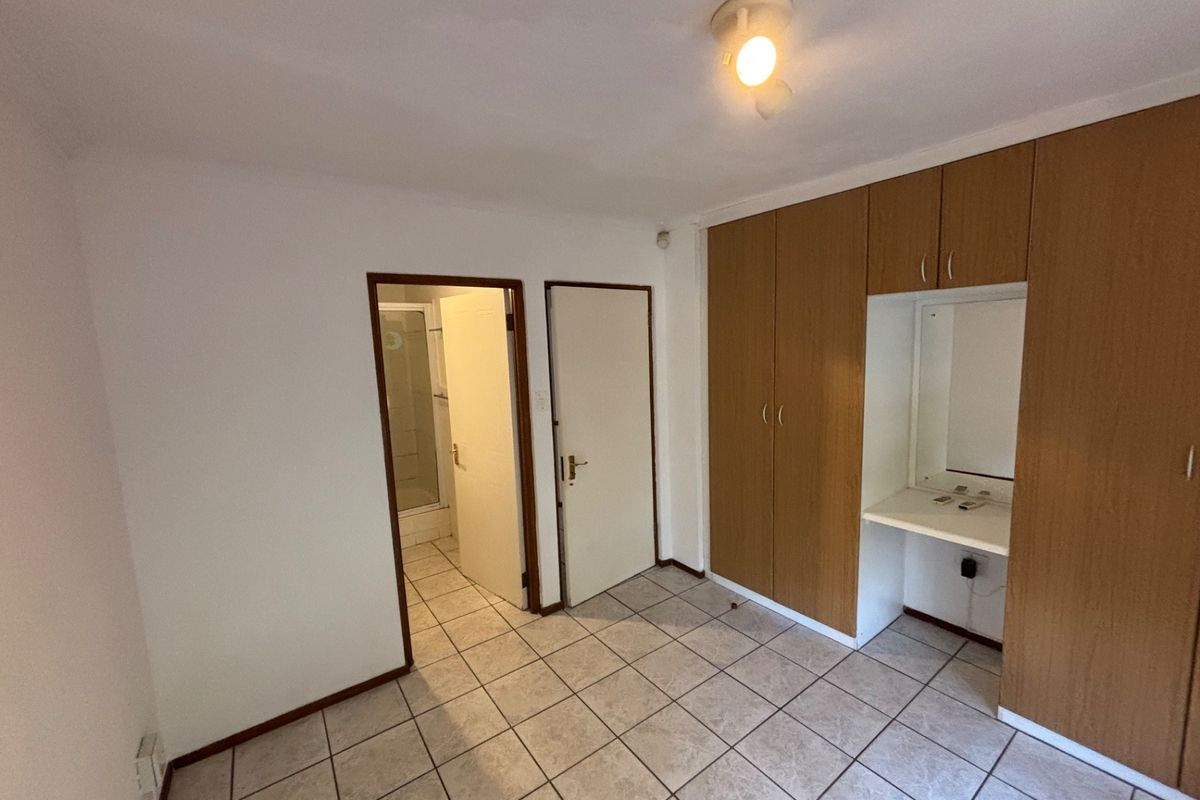 A functional bedroom featuring tiled flooring, built-in wooden cupboards with an integrated vanity area, and direct access to an en-suite bathroom.