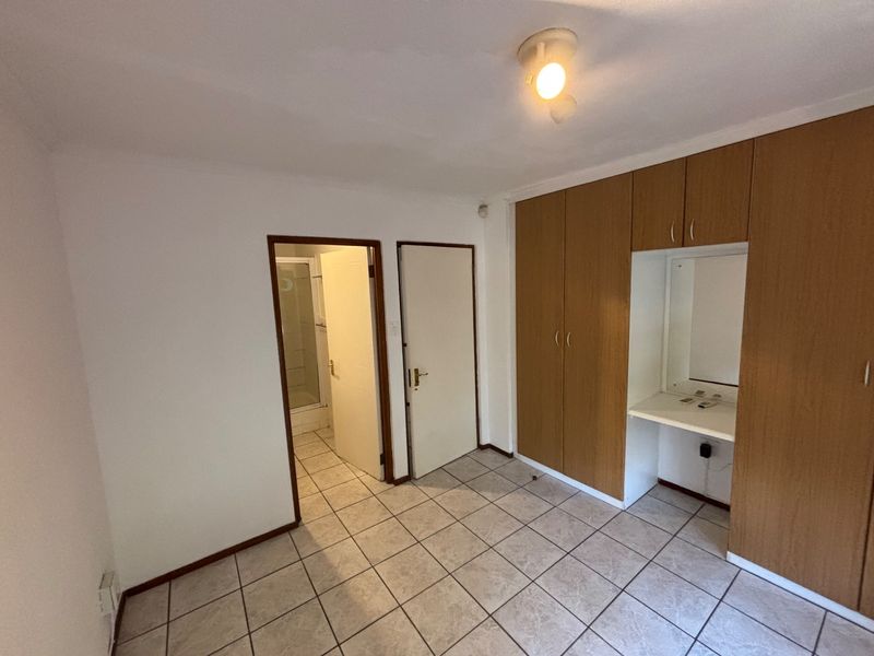A functional bedroom featuring tiled flooring, built-in wooden cupboards with an integrated vanity area, and direct access to an en-suite bathroom.