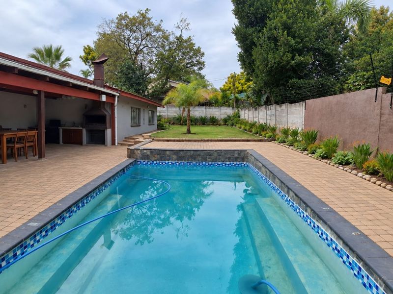 Outdoor entertainment area featuring a swimming pool, paved deck, and a covered patio with a built-in braai, set within a private garden.