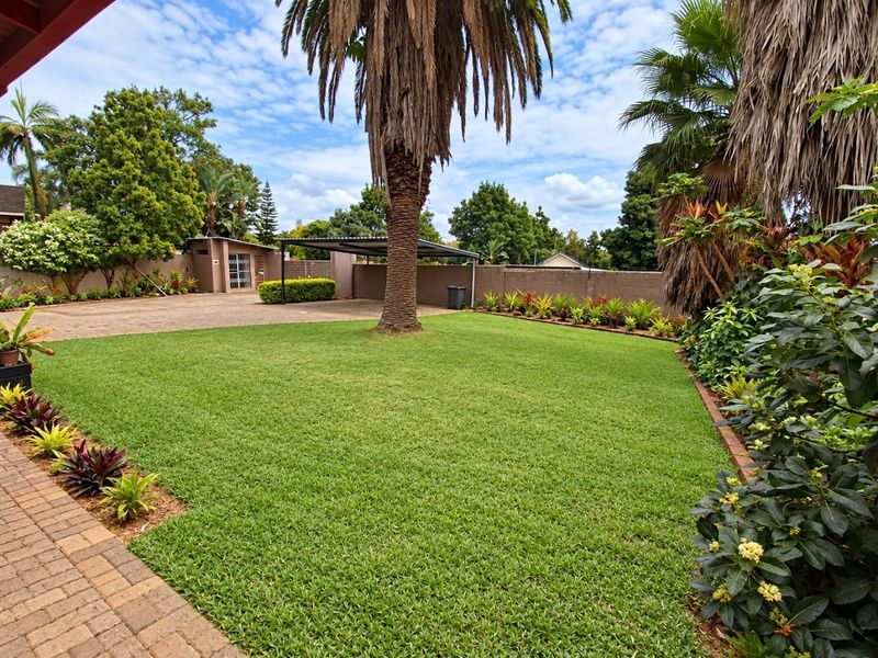 A well-maintained garden featuring a lush lawn, mature palm trees, paved driveway, and a covered carport area enclosed by a perimeter wall.