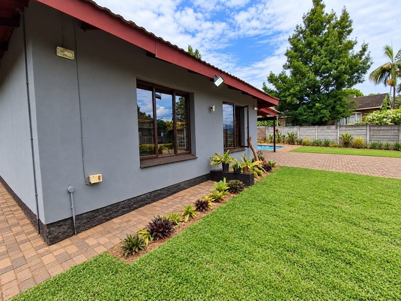 Rear exterior view of the house featuring a well-maintained lawn, brick-paved pathways, and a concrete boundary wall with a glimpse of the pool area.