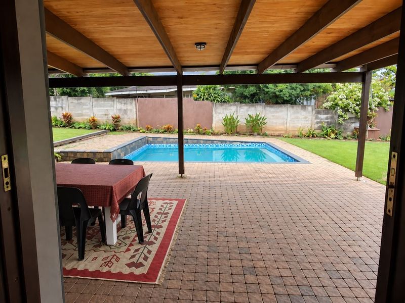 A covered outdoor patio featuring a timber-beamed ceiling, overlooking a paved pool area and a well-maintained garden with a boundary wall.