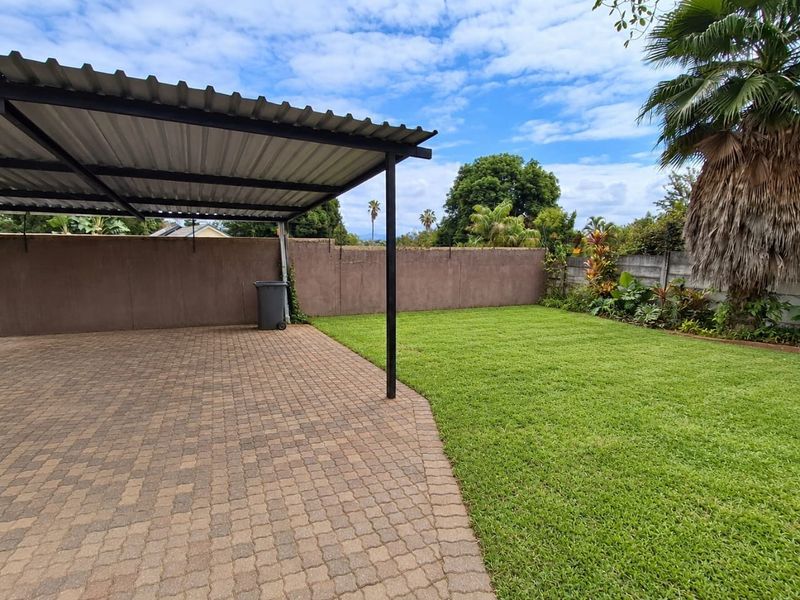 A well-maintained rear garden featuring a paved area with a covered carport, enclosed by a solid boundary wall and established lawn.
