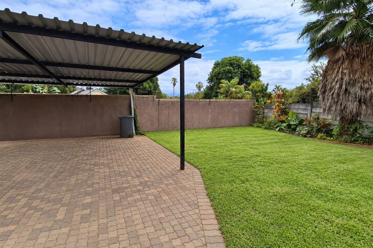 A well-maintained rear garden featuring a paved area with a covered carport, enclosed by a solid boundary wall and established lawn.