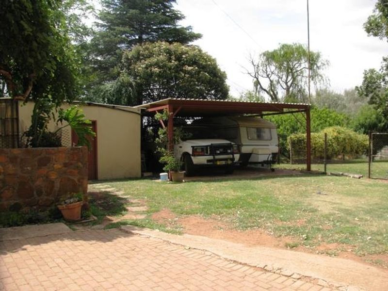 A rear exterior view of the property featuring a covered carport structure, stone walling, and paved driveway area set within a garden.