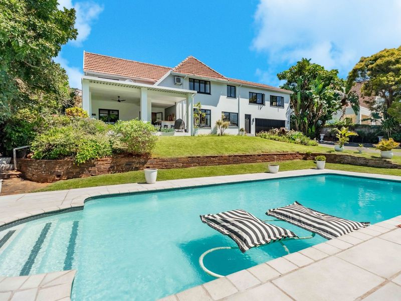 A well-maintained rear garden featuring a swimming pool, stone retaining wall, and a spacious covered patio attached to the main residence.